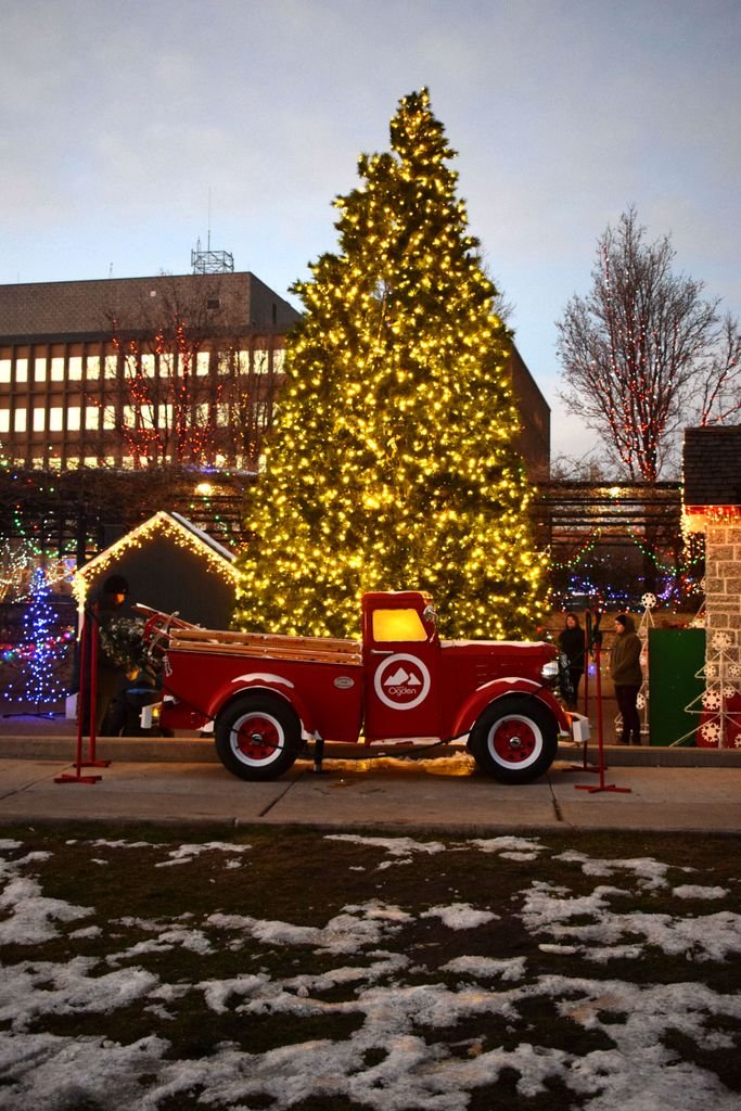 A red truck is parked in front of a brightly lit Christmas tree in a festive outdoor setting at dusk.